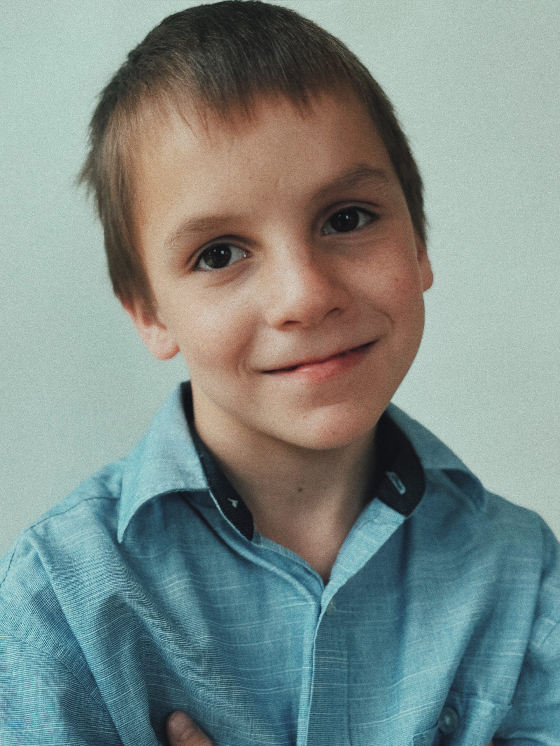 Child in a blue collared shirt smiling at the camera against a pale background.