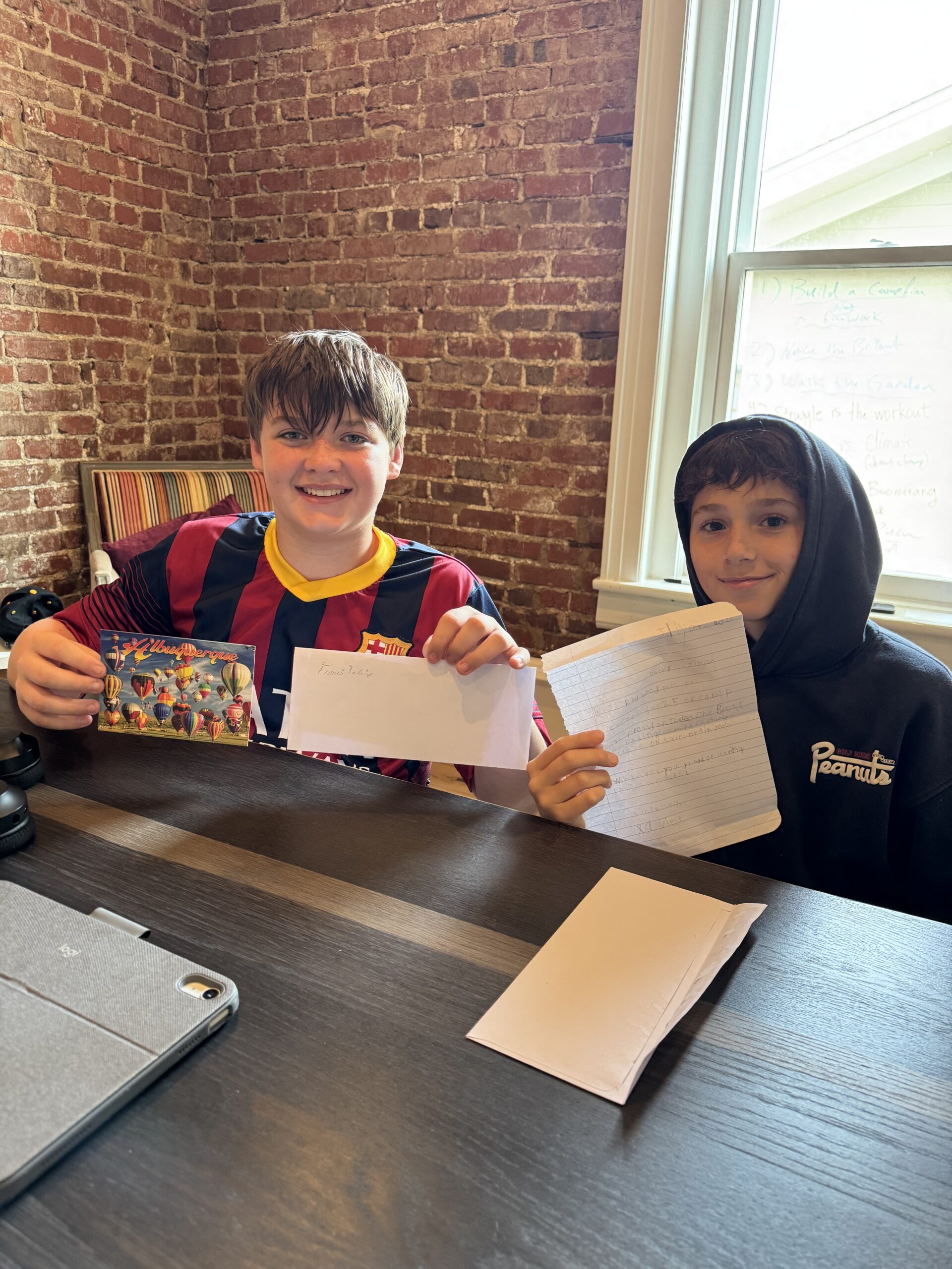 Two smiling students proudly holding up their written work and a postcard at a table at Redwood in Nasvhille.